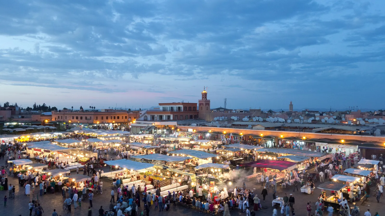 Place Jemaa el-Fna au crépuscule, stands illuminés et foule, minaret de la Koutoubia au loin